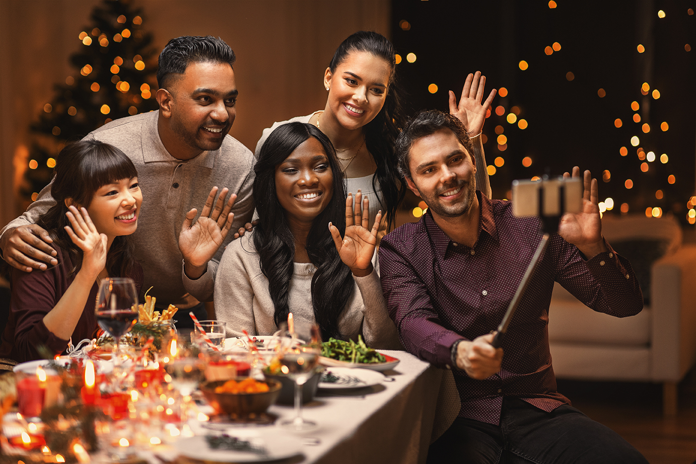 A group of friends around a table taking a photo with a selfie stick. The room is decorated for the winter holiday season.